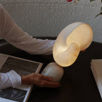 A hand tilting the conch shade of this Clérachic alabaster lamp on its stone base, set on a dark table beside open magazines.