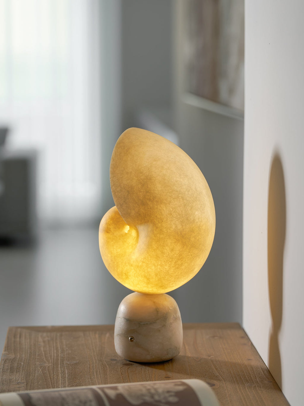 Clérachic table lamp with a glowing alabaster conch shade on a marbled stone base, standing on a wooden table in a bright room.
