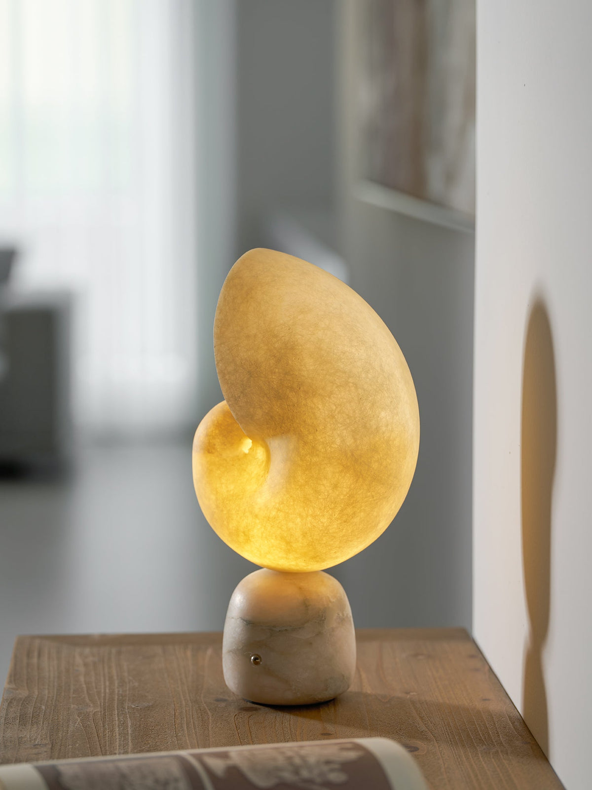 Clérachic table lamp with a glowing alabaster conch shade on a marbled stone base, standing on a wooden table in a bright room.
