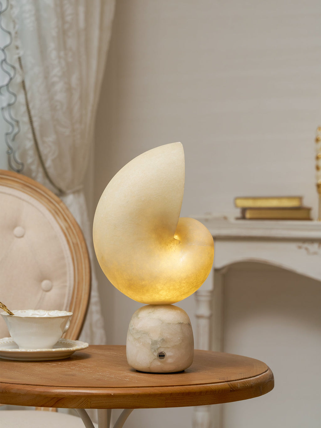 This Clérachic alabaster lamp on a round wooden table beside a teacup, with a cane-backed chair and a lace curtain behind.