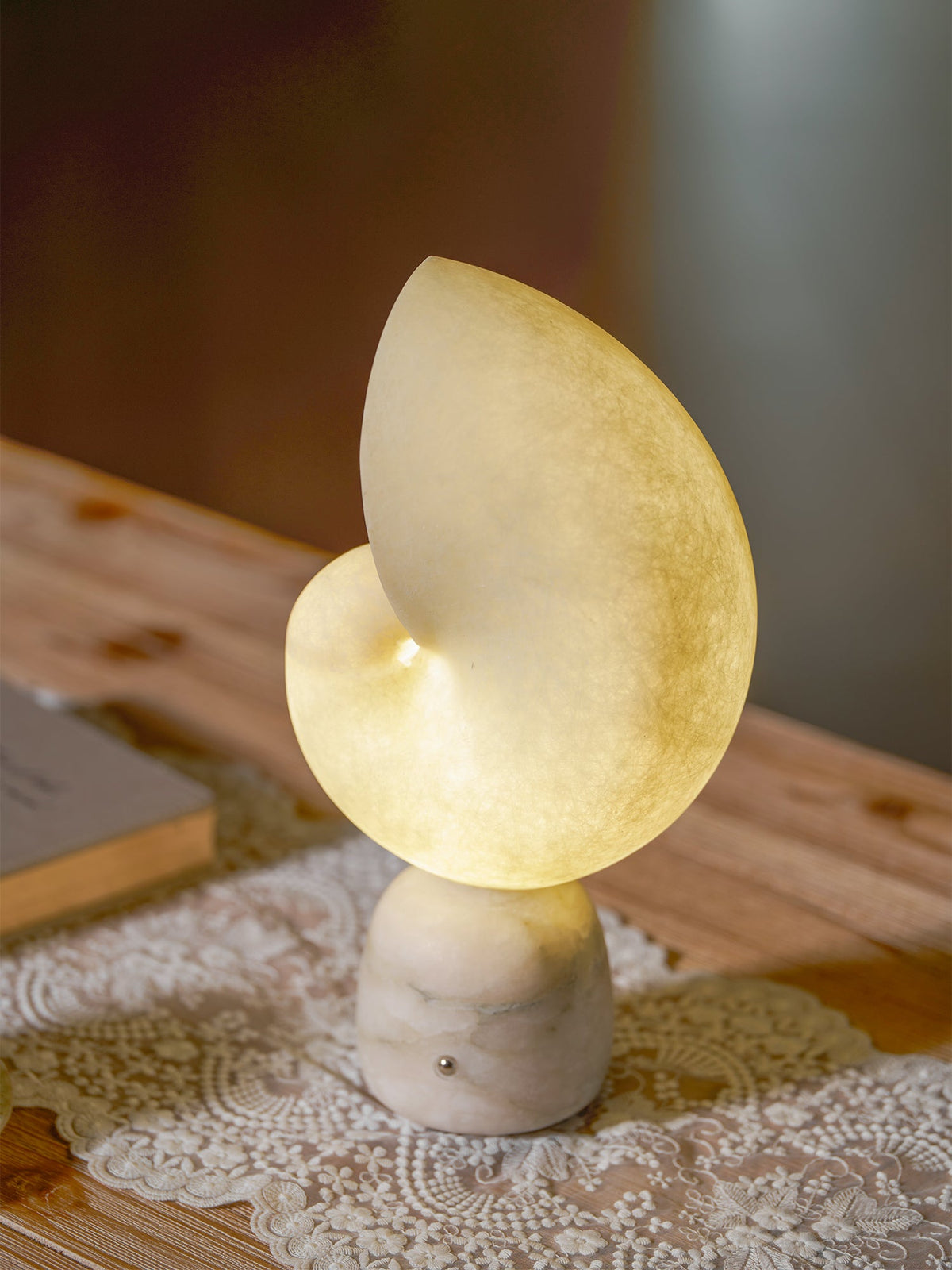 Angled view of a lit Clérachic conch lamp on a lace runner over a wooden table, with a firelit room blurred behind.