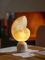 Clérachic alabaster conch lamp glowing amber on a lace runner, with a book on the table and a fireplace blurred in the background.