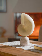 Unlit Clérachic conch lamp in pale cream alabaster on a lace table runner, with a book to one side and a fireplace behind.