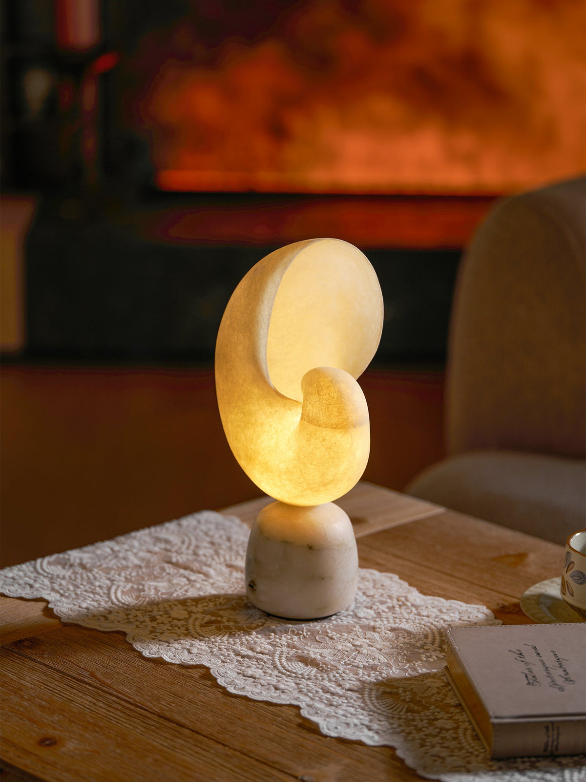 A smaller view of the lit Clérachic alabaster conch lamp on a lace runner in a living room, with an armchair and a fire glowing behind.