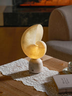 Clérachic table lamp with a softly lit alabaster conch shade on a lace runner, with an armchair and a closed book alongside.