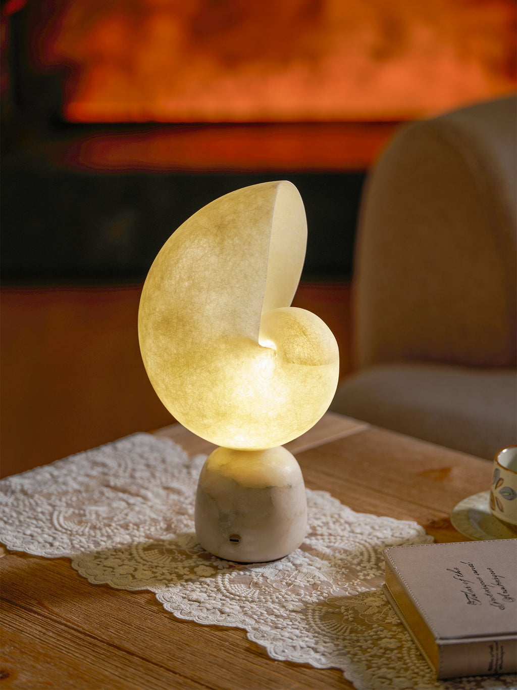 Lit Clérachic conch lamp on a lace runner beside a book and a cup, with an upholstered chair and a warm fire blurred behind.