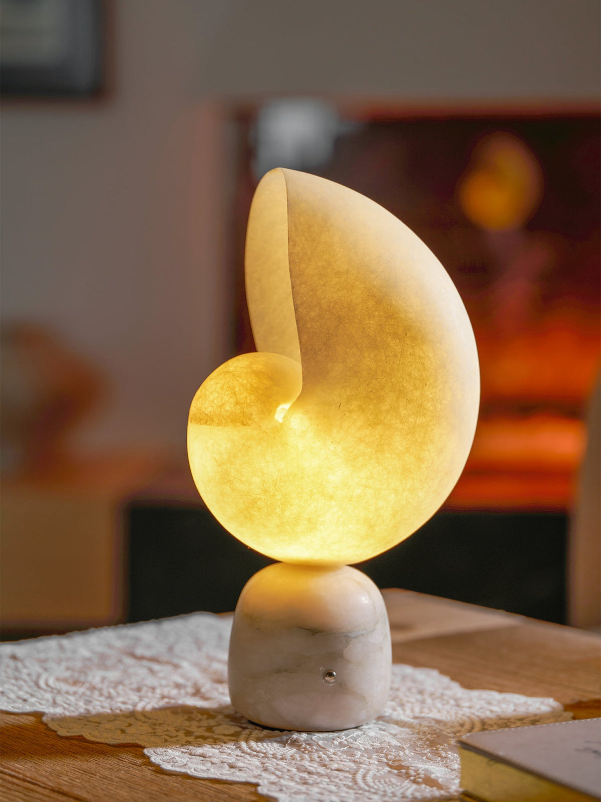 Close view of a lit Clérachic alabaster conch lamp with a golden glow, standing on a lace runner on a wooden table.