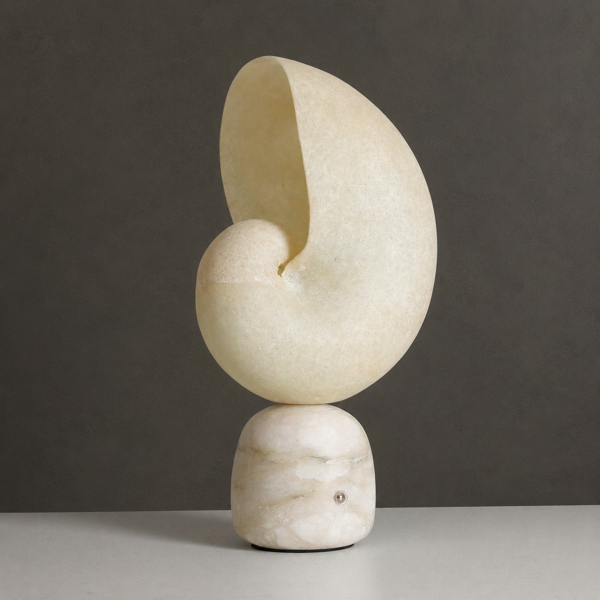 Unlit Clérachic alabaster conch lamp on a marbled stone base with a small metal touch point, against a dark grey backdrop.