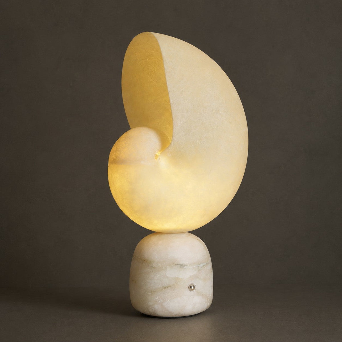 The same Clérachic alabaster lamp lit, the conch shade glowing warm yellow against a dark grey backdrop.