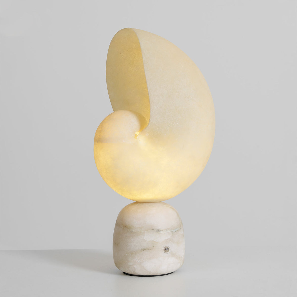 Lit Clérachic alabaster lamp seen at a slight angle, the conch shade glowing warm over its white veined stone base on pale grey.
