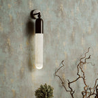Black Clérachic Alabaster Tube Wall Sconce lit on a weathered green plaster wall above dried branches in a quiet corner.