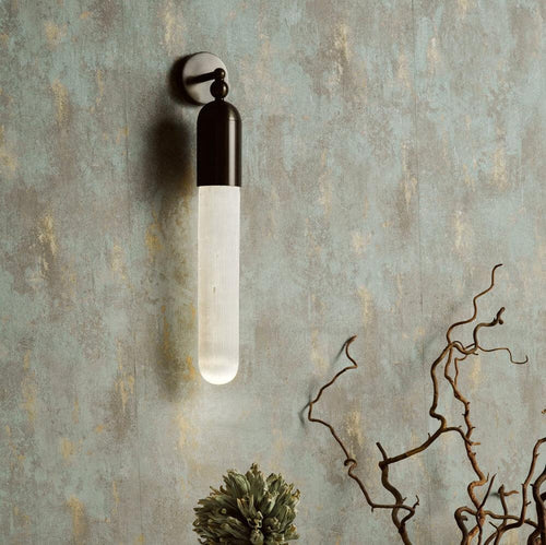 Black Clérachic Alabaster Tube Wall Sconce lit on a weathered green plaster wall above dried branches in a quiet corner.