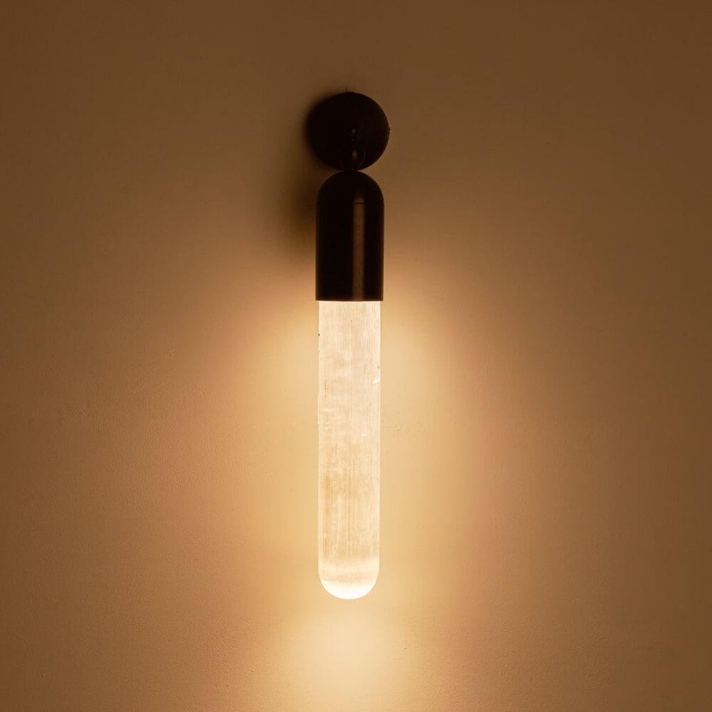 Clérachic Alabaster Tube Wall Sconce switched on in a dark room, the black cap above a stone tube glowing warm on the wall.