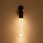 Clérachic Alabaster Tube Wall Sconce switched on in a dark room, the black cap above a stone tube glowing warm on the wall.