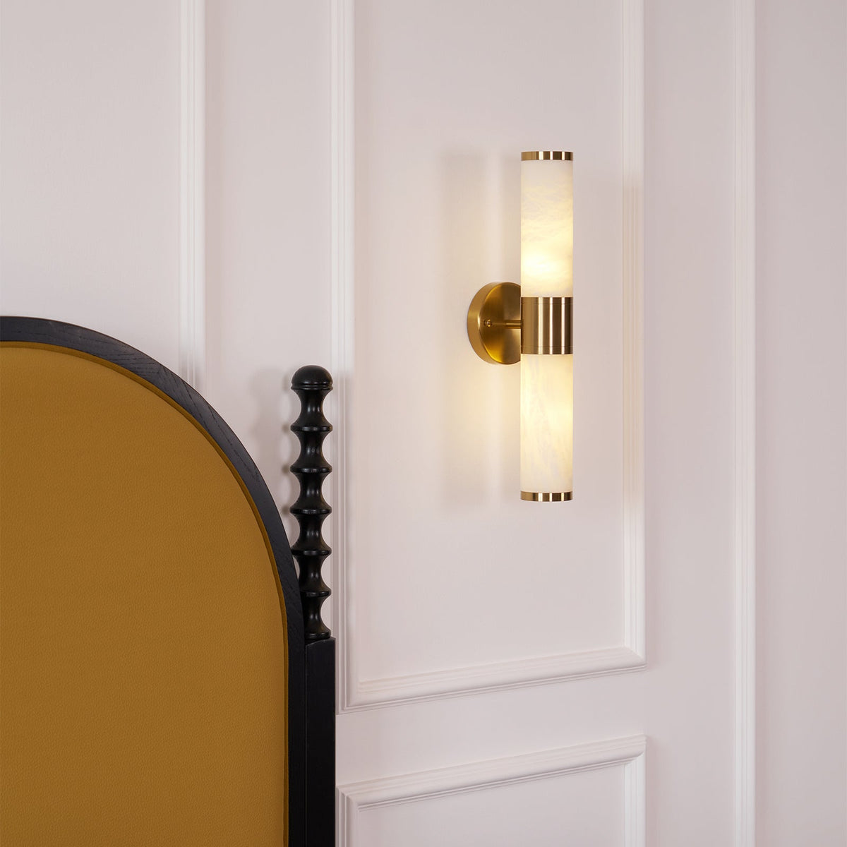 Lit Clérachic wall light with veined white tubes on a panelled white wall, beside an arched mustard-velvet headboard with a black turned post.