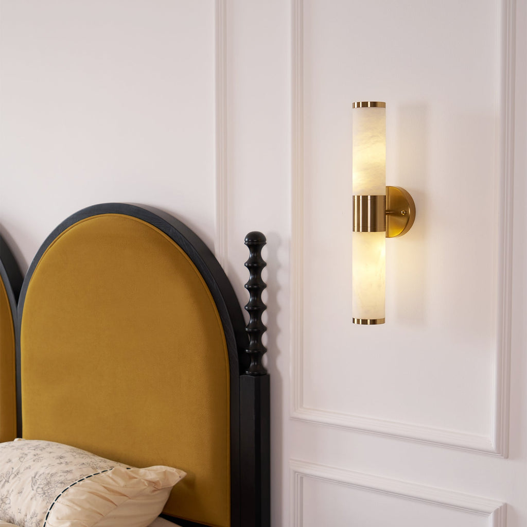 A Clérachic wall light glowing in two halves on a white panelled wall above the arched mustard headboard of a bed with floral bedding.