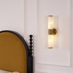Clérachic twin-tube wall light with a gold centre band, lit above a black bedside post and a panelled white wall.
