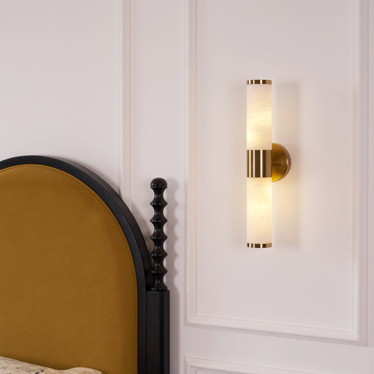 Clérachic twin-tube wall light with a gold centre band, lit above a black bedside post and a panelled white wall.