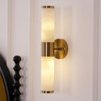 Close view of the lit alabaster tubes and gold band on this Clérachic wall light, with the black turned bedpost at the left edge.