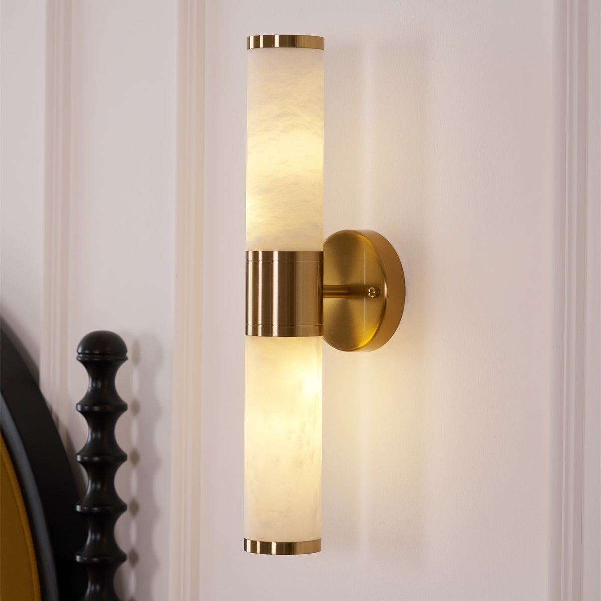Close view of the lit alabaster tubes and gold band on this Clérachic wall light, with the black turned bedpost at the left edge.