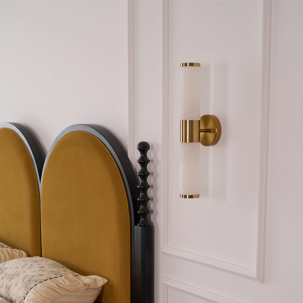 This Clérachic wall light, switched off, shows pale veined tubes and a gold band beside two arched mustard headboards and a black bedpost.