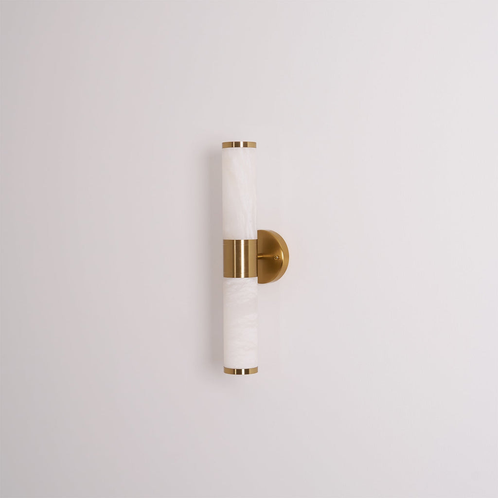 Unlit veined white tubes with gold end caps and a round gold plate, this Clérachic wall light on a plain pale grey wall.