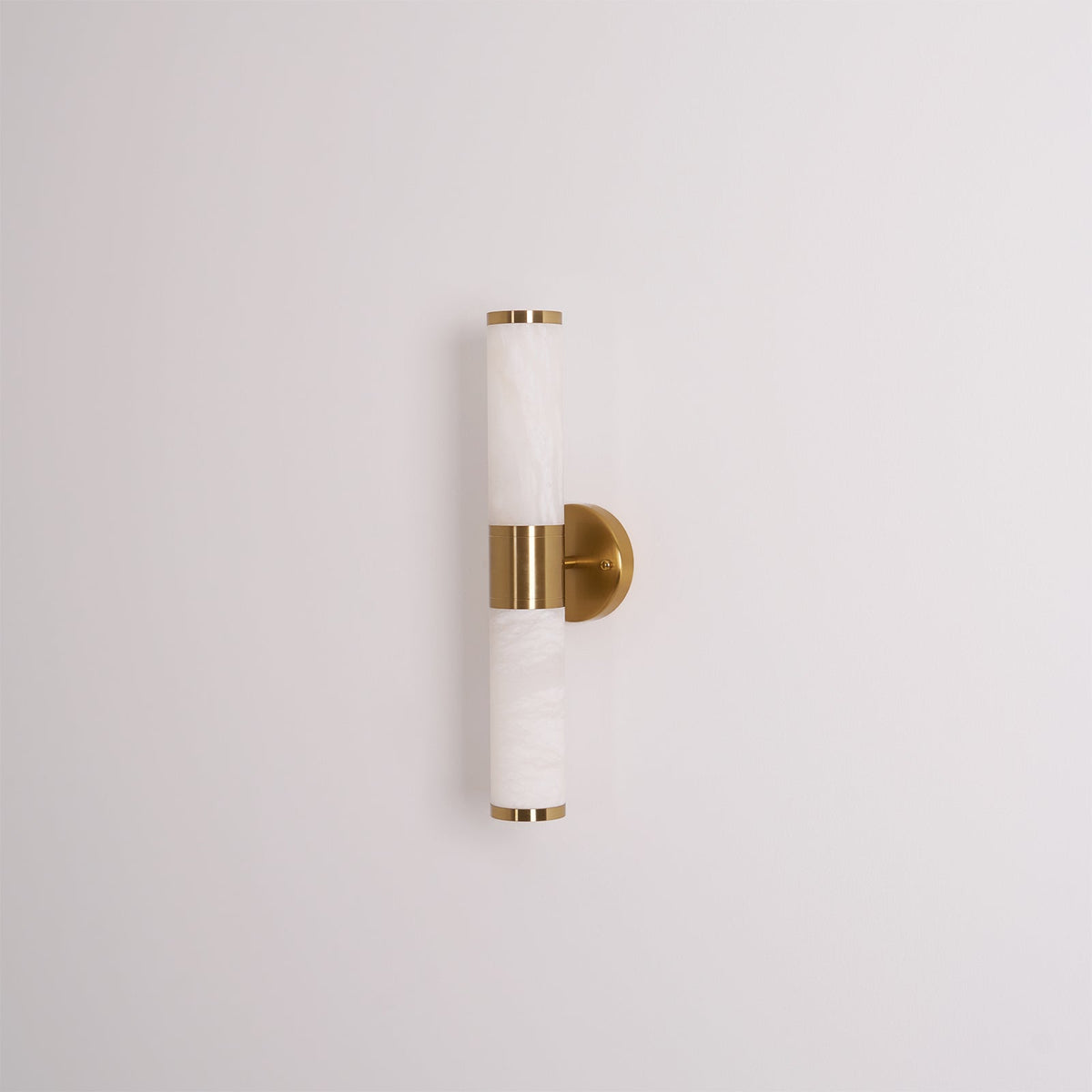 Unlit veined white tubes with gold end caps and a round gold plate, this Clérachic wall light on a plain pale grey wall.