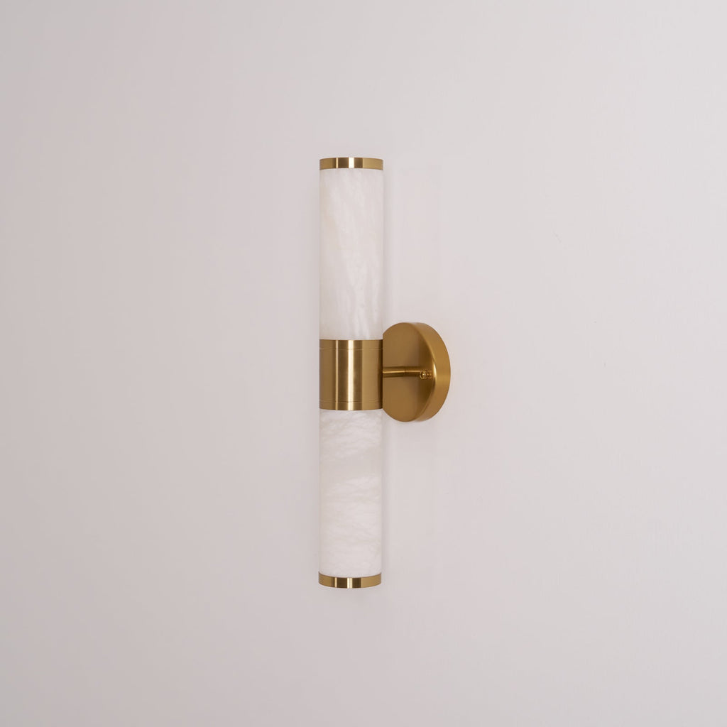 Straight-on view of this Clérachic wall light with its veined white tubes, gold caps and round gold plate on a pale wall, switched off.