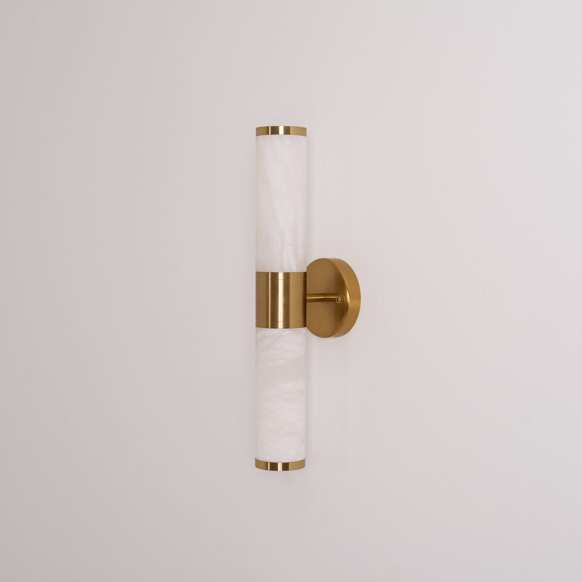 Straight-on view of this Clérachic wall light with its veined white tubes, gold caps and round gold plate on a pale wall, switched off.