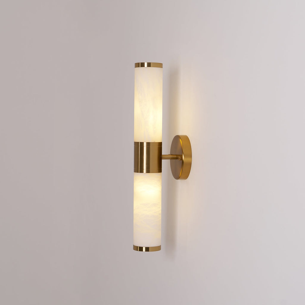 Clérachic wall light lit against a mid-grey wall, with a warm glow in the lower tube and a gold band at the centre.