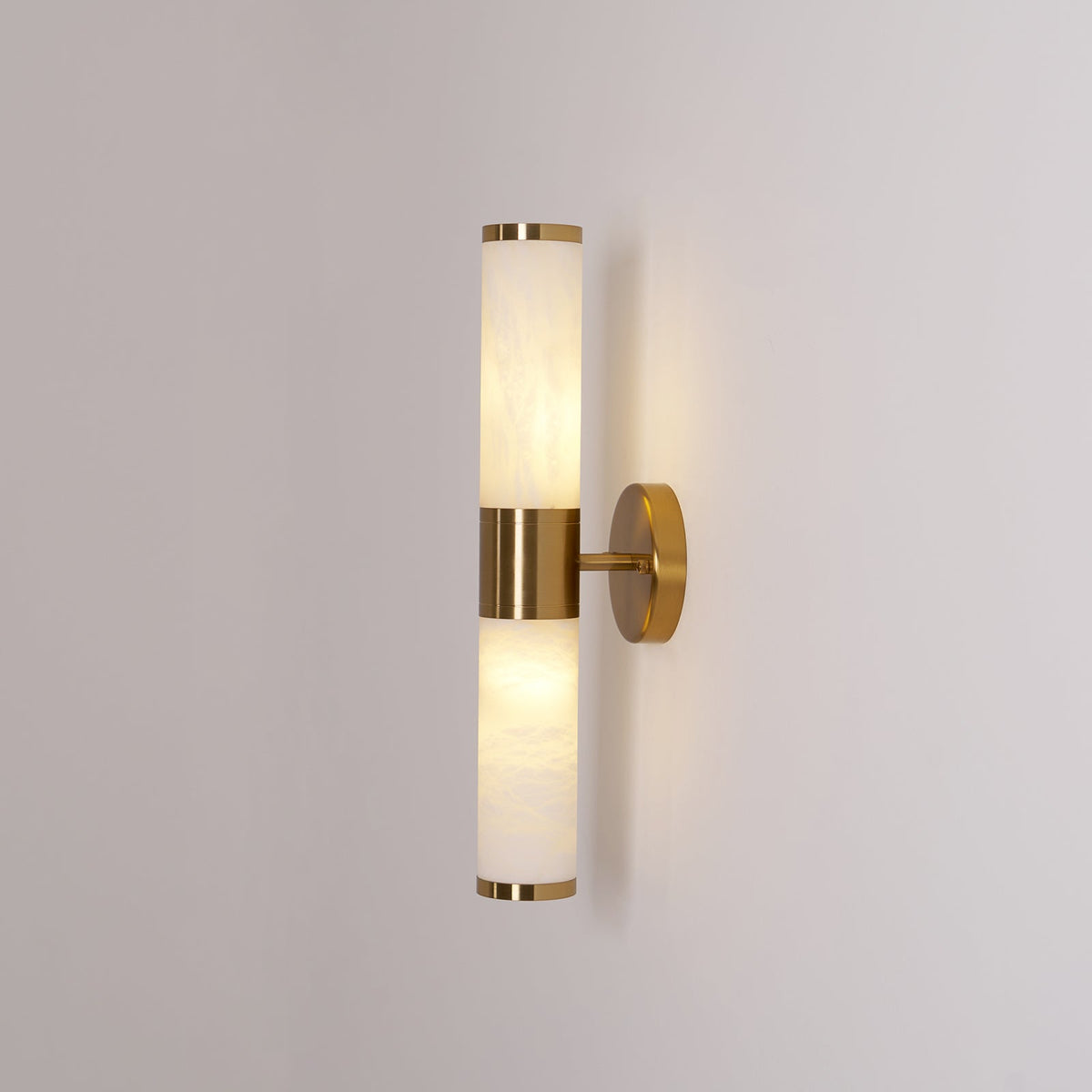 Clérachic wall light lit against a mid-grey wall, with a warm glow in the lower tube and a gold band at the centre.