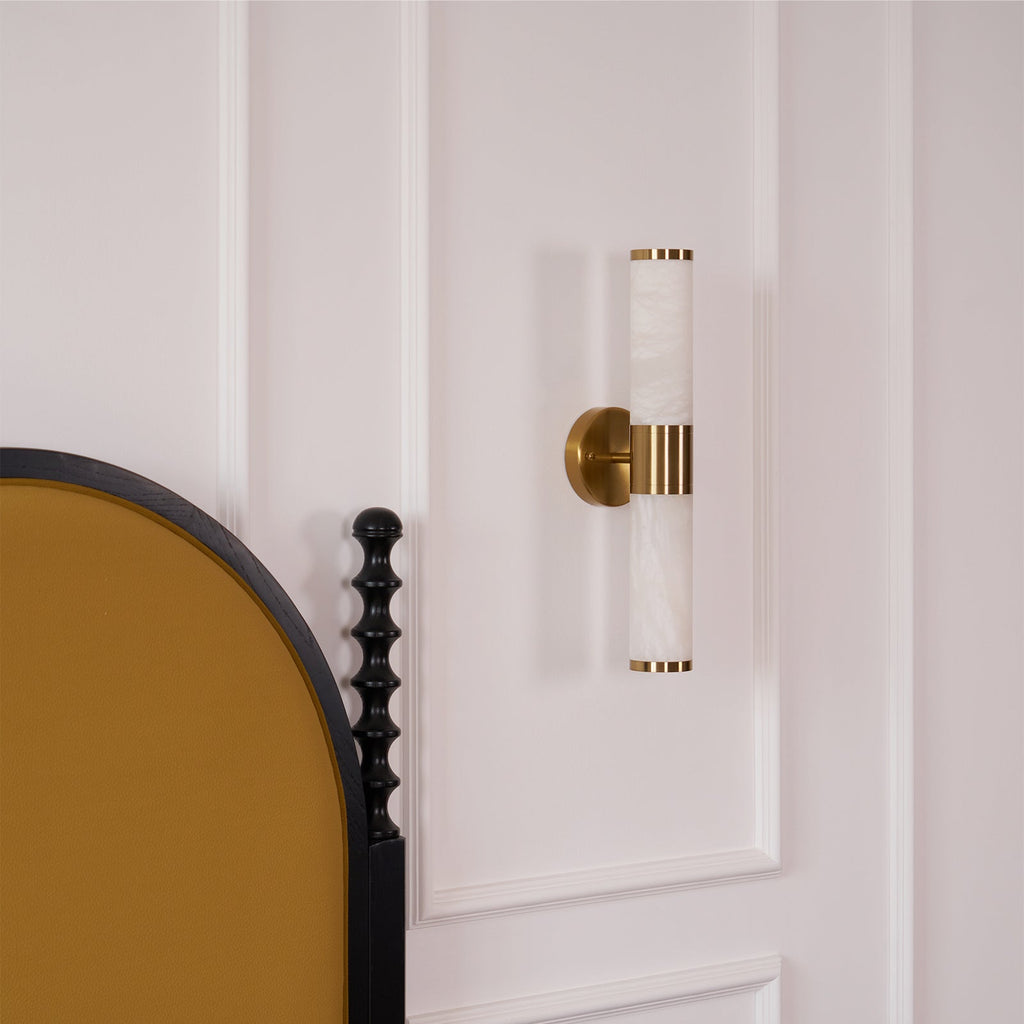Small view of the Clérachic wall light on a white panelled wall beside an arched mustard headboard and a black turned bedpost.