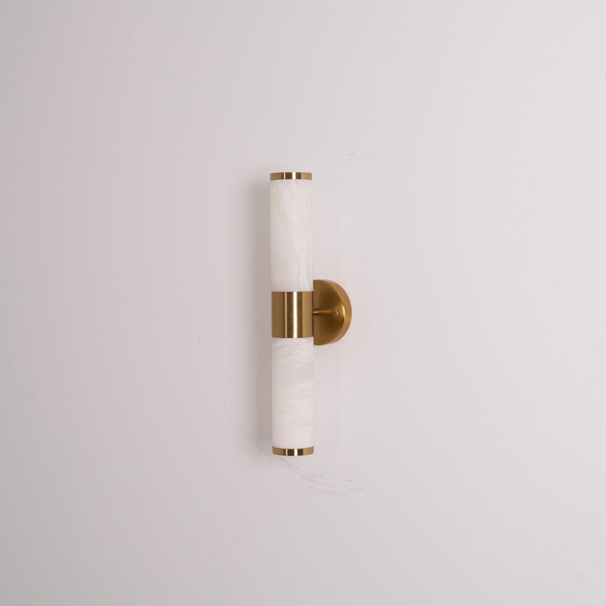 Unlit Clérachic wall light on a pale wall, with veined white tubes, a gold band and a round gold plate seen from a slight angle.