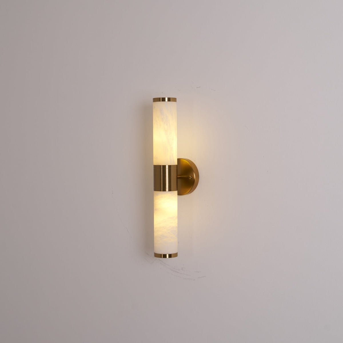 Lit Clérachic wall light on a grey wall, with warm light inside the veined tubes and a gold collar and wall plate.