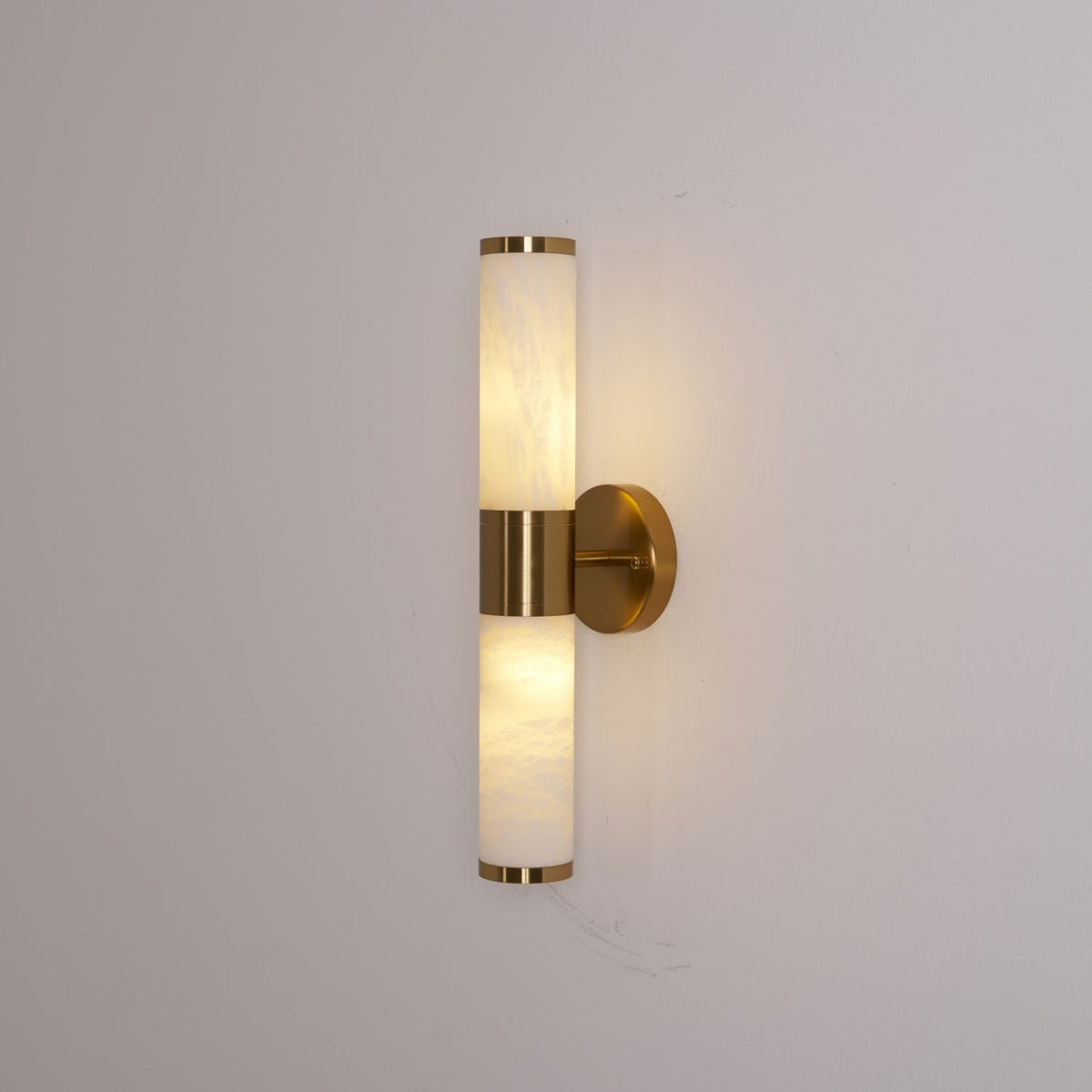 A Clérachic wall light glowing evenly in both veined white tubes against a soft grey wall, with a gold collar at the centre.