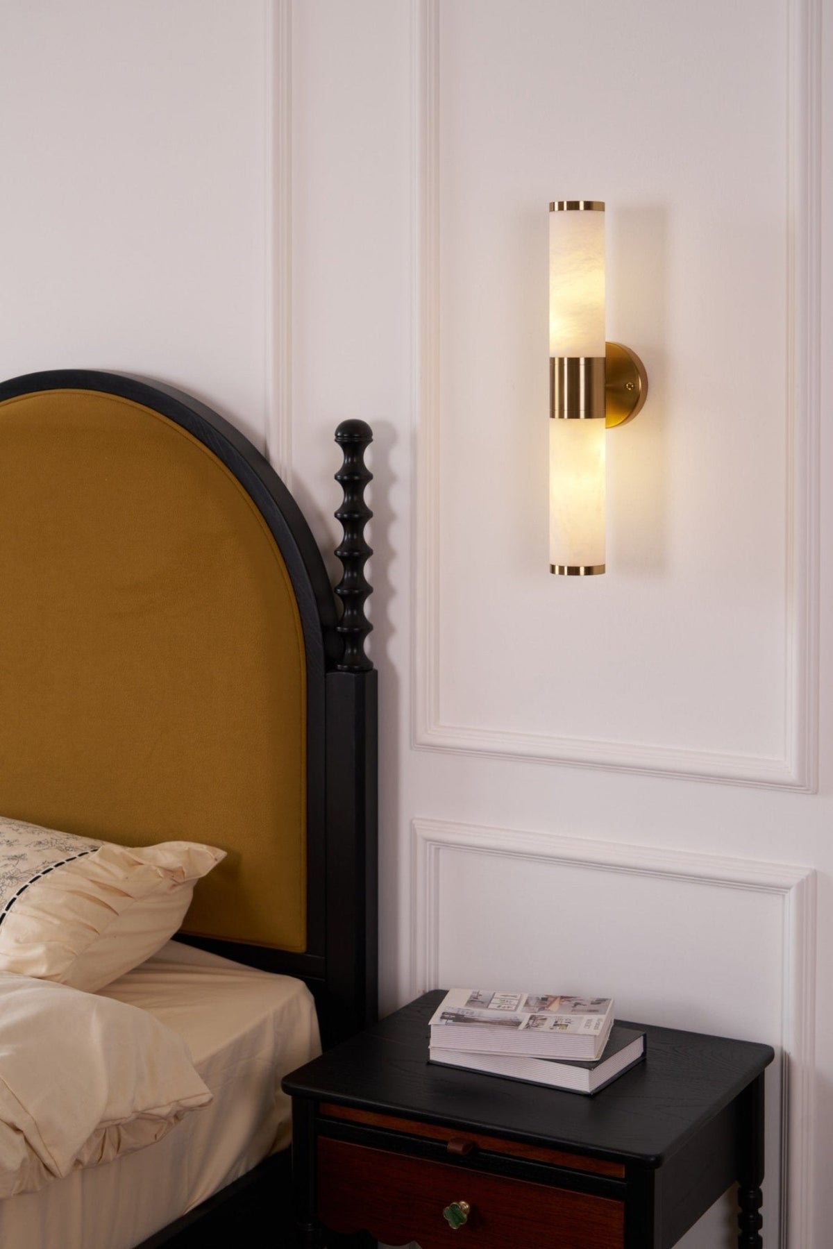 Lit Clérachic wall light on a panelled wall above a black bedside table with books, beside an arched mustard headboard and cream bedding.