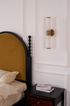 Unlit Clérachic wall light on a white panelled wall, above a dark bedside table with books and a mustard arched headboard.