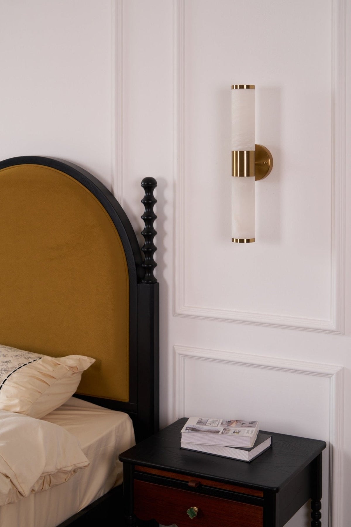 Unlit Clérachic wall light on a white panelled wall, above a dark bedside table with books and a mustard arched headboard.