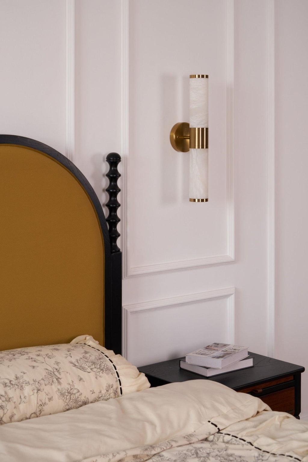 This Clérachic wall light, switched off, hangs on a panelled wall above a black nightstand and a bed with floral cream bedding.