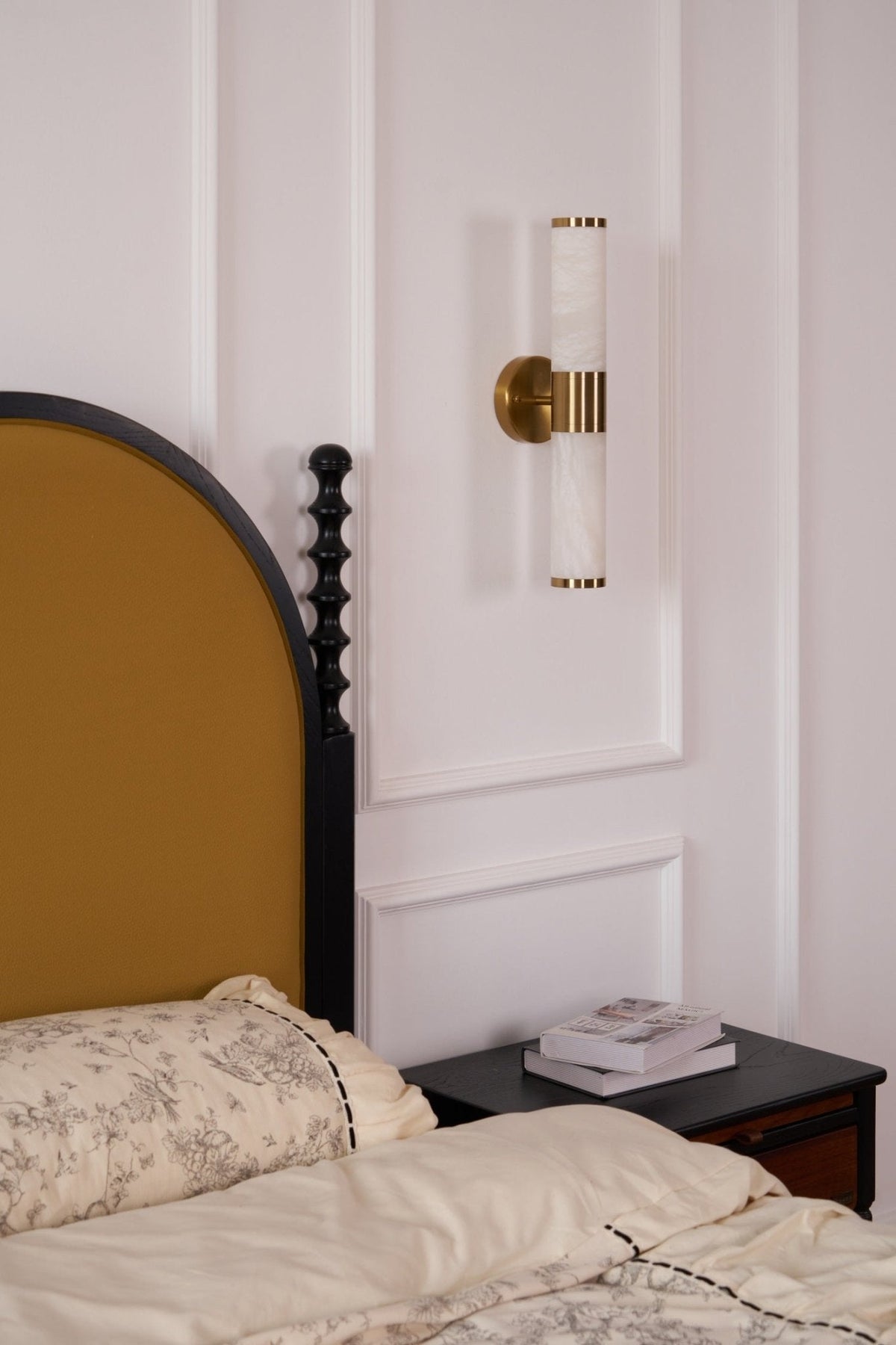 This Clérachic wall light, switched off, hangs on a panelled wall above a black nightstand and a bed with floral cream bedding.