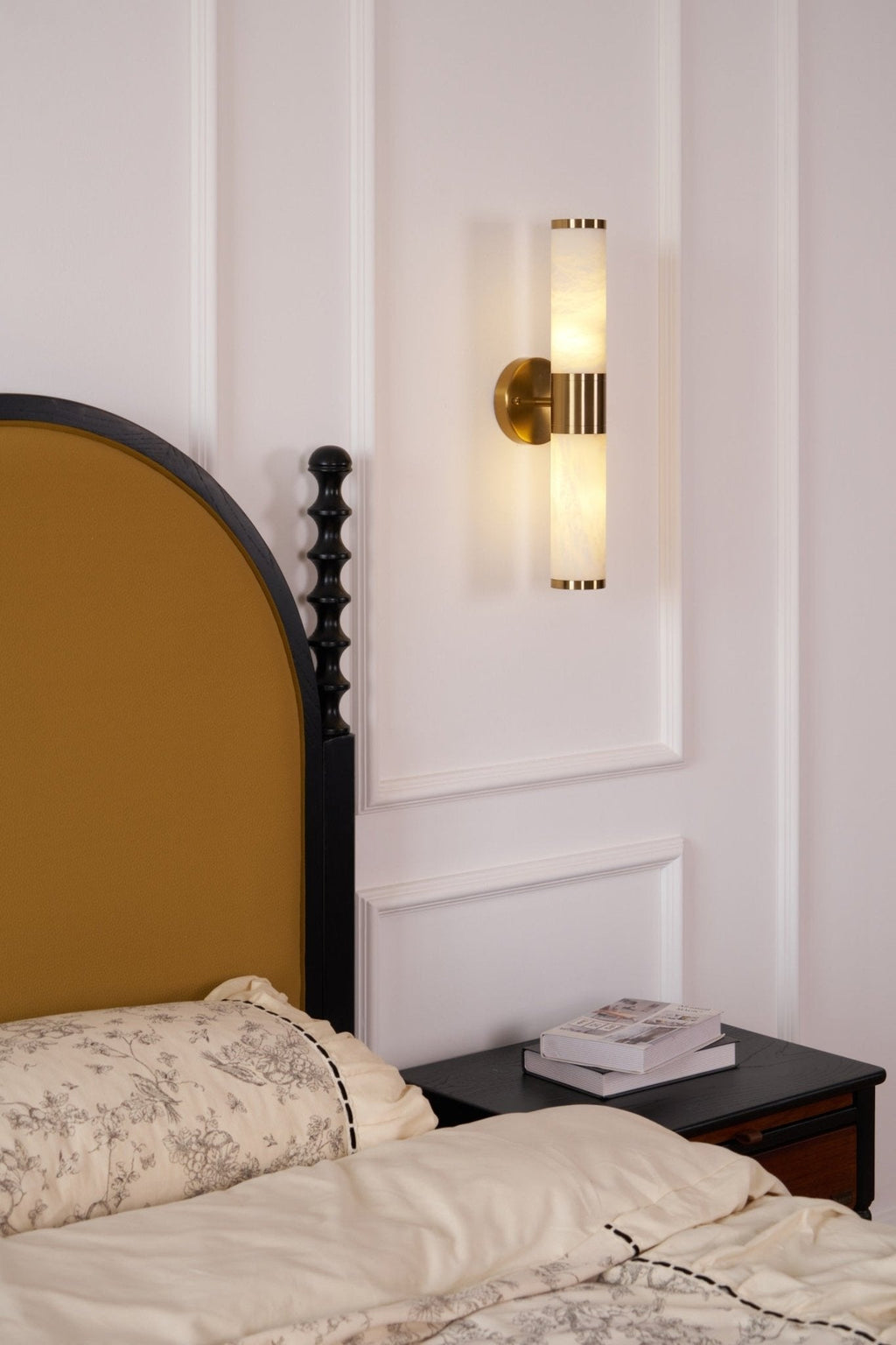 Clérachic wall light glowing warmly above the foot of a bed with floral cream bedding and an arched mustard headboard.
