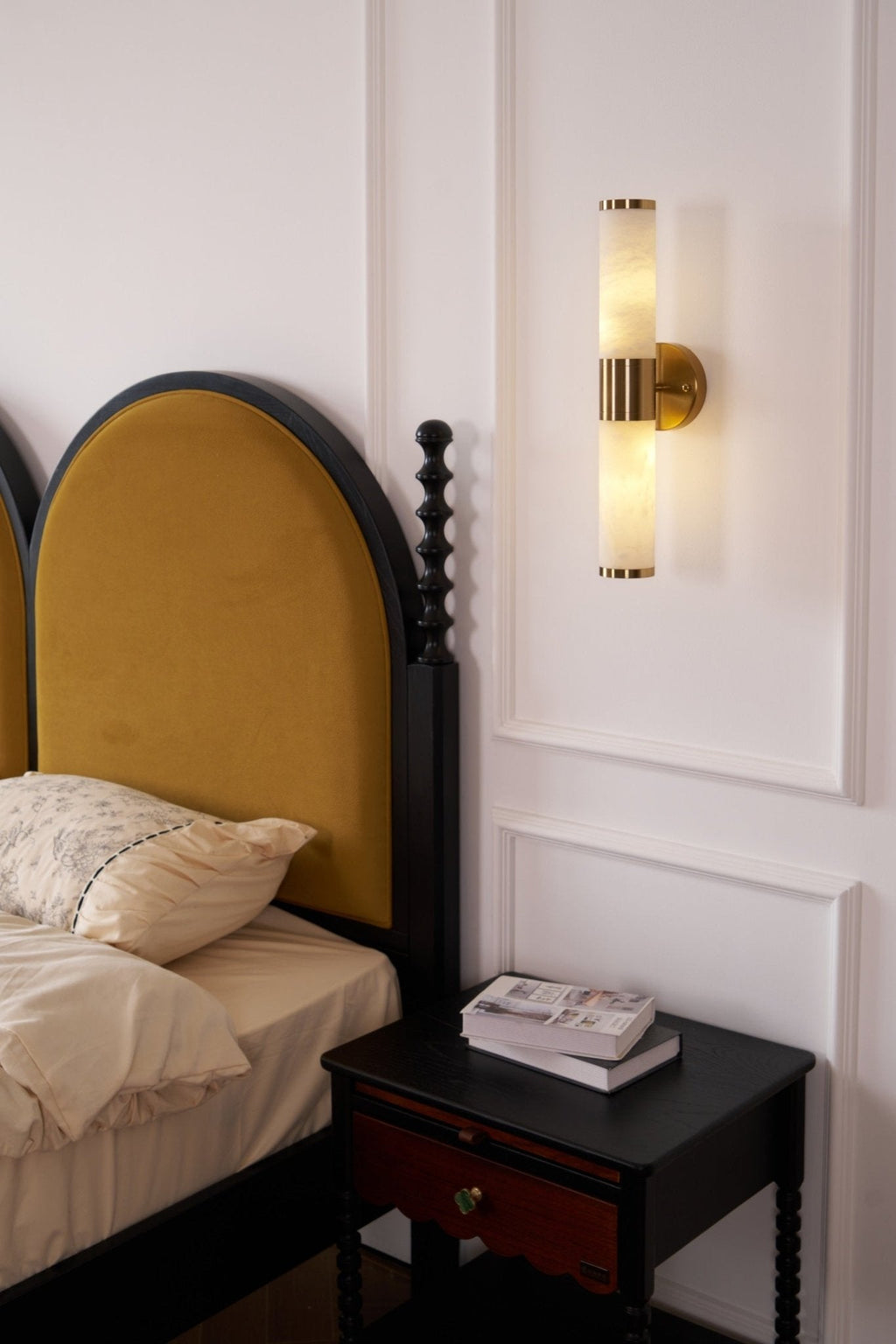 A lit Clérachic wall light beside a mustard arched headboard and a black nightstand with a stack of books on a panelled wall.