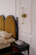 Switched-off Clérachic wall light on a white panelled wall, with two arched mustard headboards and a black nightstand below.