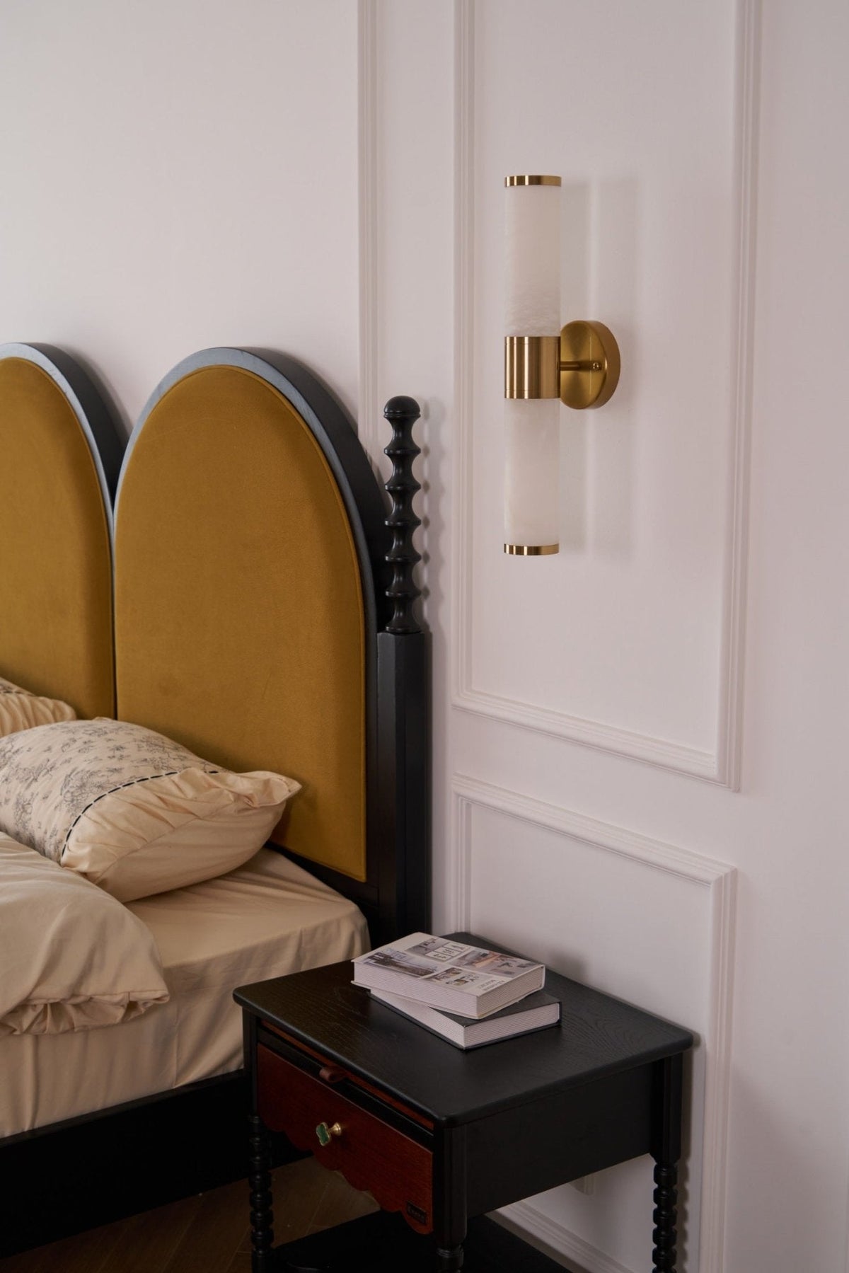 Switched-off Clérachic wall light on a white panelled wall, with two arched mustard headboards and a black nightstand below.