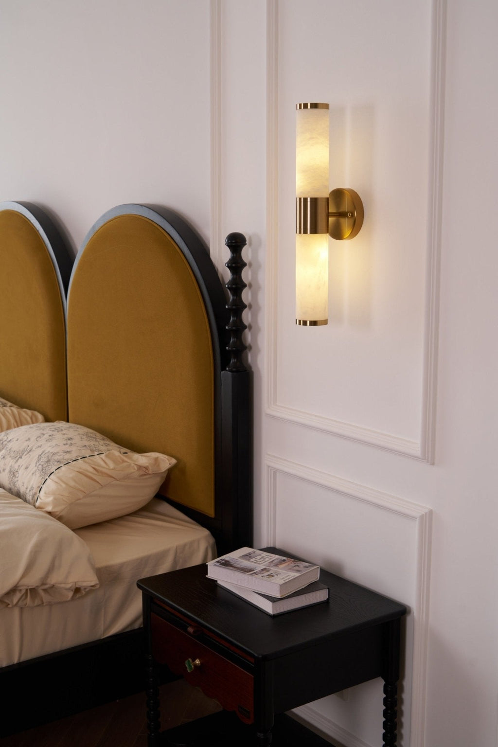 Lit Clérachic wall light with two veined tubes above a dark nightstand and twin arched mustard headboards in a panelled bedroom.