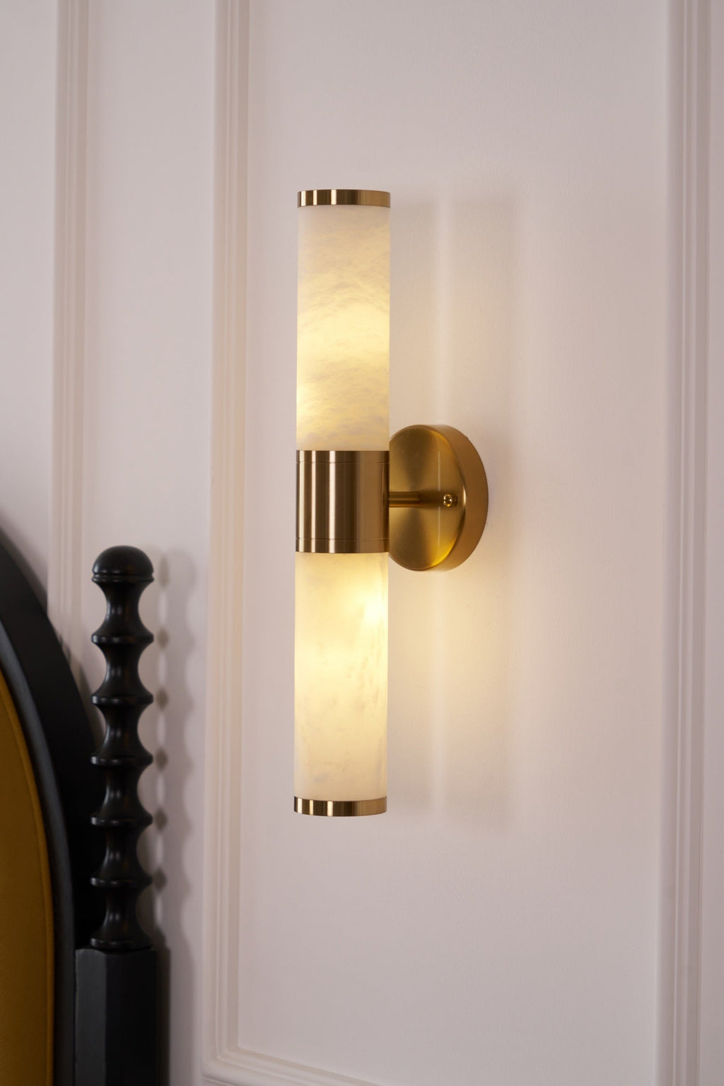 Close view of the lit veined alabaster tubes and gold band of this Clérachic wall light, with a black turned bedpost at the lower left.