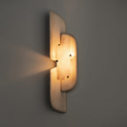 A Clérachic wall lamp glowing amber on a dark grey wall, with a fan of light escaping to the left of its beige panels.