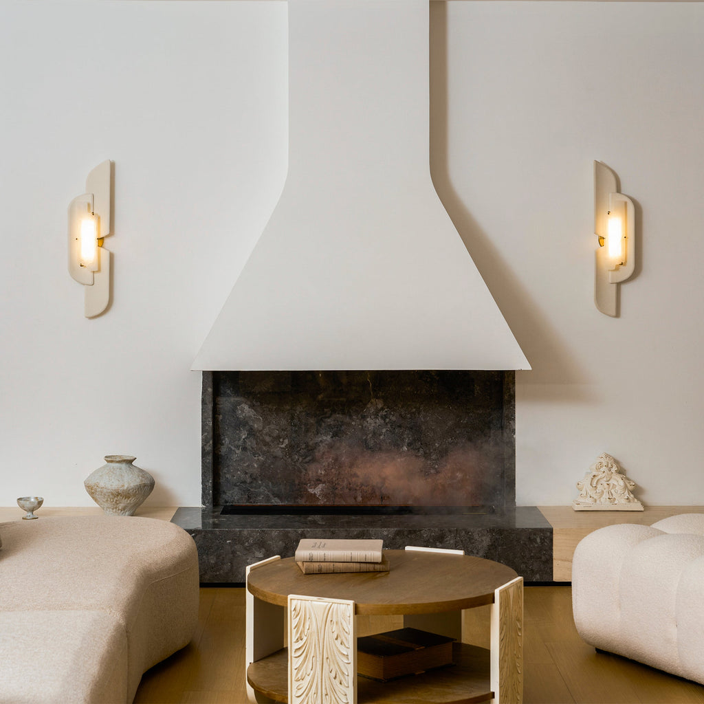 A pair of Clérachic wall lamps flank a plaster fireplace hood above round tables and curved cream sofas, both softly lit.
