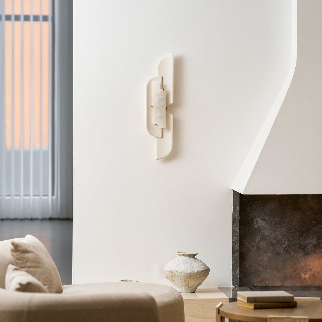 A Clérachic wall lamp on a curved white plaster wall, with a stone fireplace and a textured vase at the bottom of the frame.