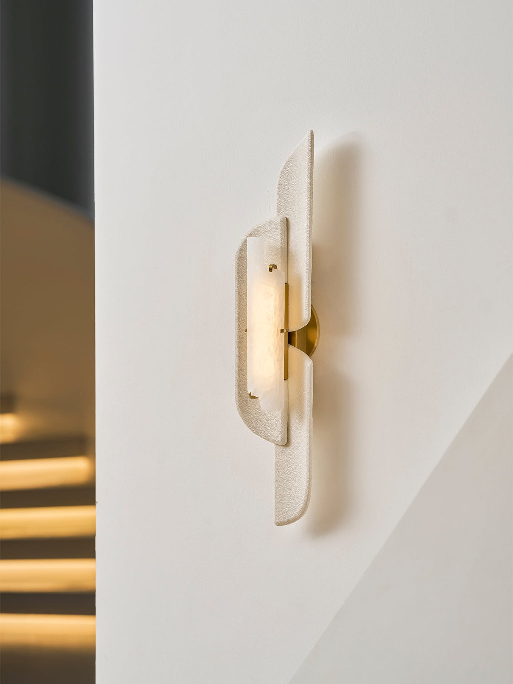 A Clérachic wall lamp seen at an angle on a white wall, a brass bracket showing between its panels, with a lit stair behind.